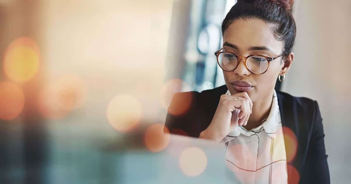 A woman with glasses and a bun looks intently at a laptop screen, wearing business attire in an office setting with blurred lights in the foreground, focused on senior living sales strategies.
