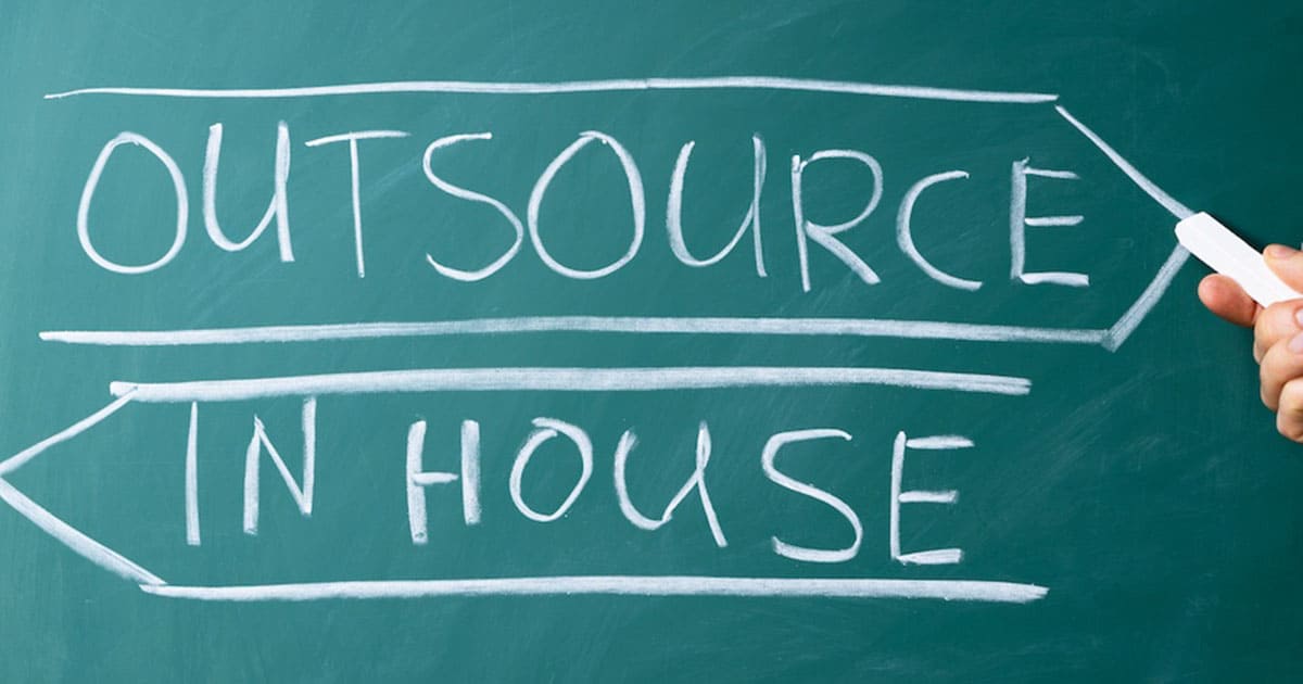 A hand writes the words "OUTSOURCE" and "IN HOUSE," with arrows pointing in opposite directions on a green chalkboard, illustrating the choices for managing a senior living call center.