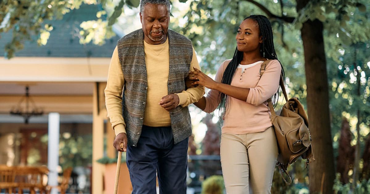 An older man walks with a cane outdoors while a younger woman, perhaps inspired by senior living marketing tips, walks beside him, holding his arm supportively.