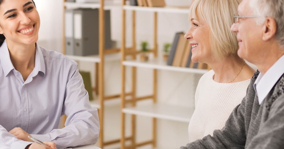 Three people, two older adults and a younger professional, sit at a table discussing customer experience marketing in an office setting with shelves in the background.