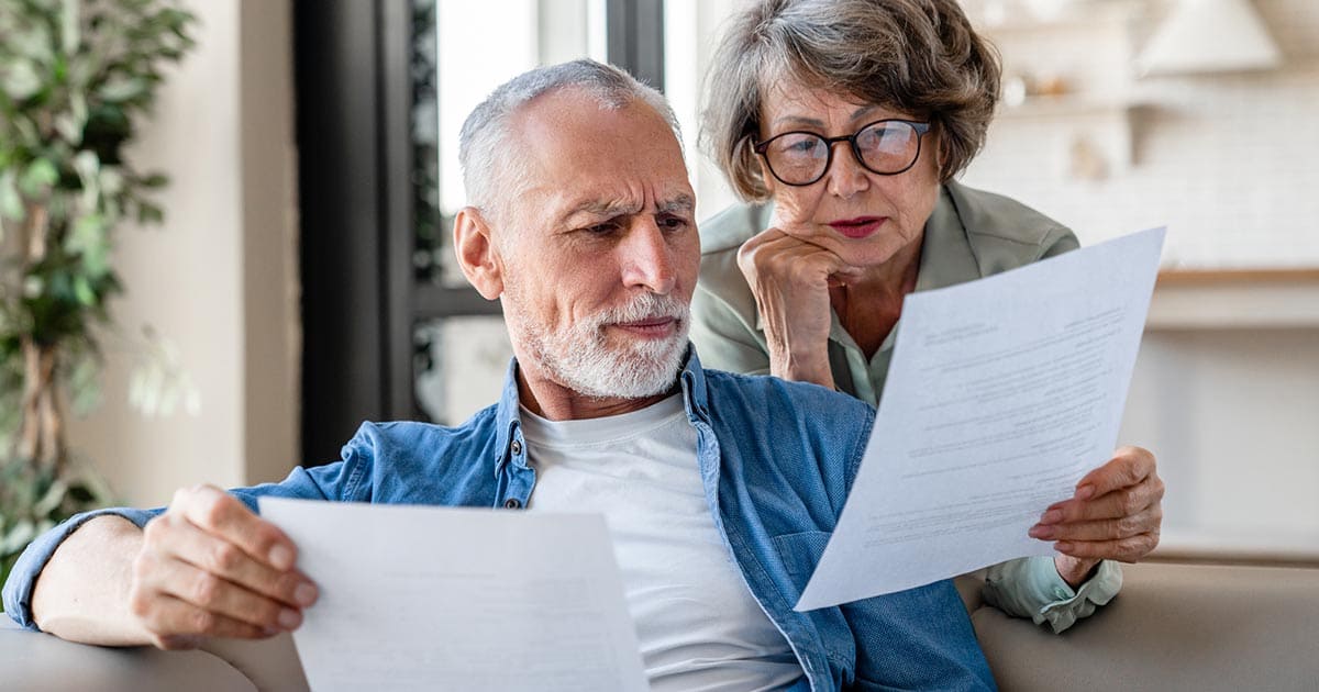 An older man and woman sit on a couch in a well-lit room, closely reading and examining papers together as they discuss senior living sales tips.