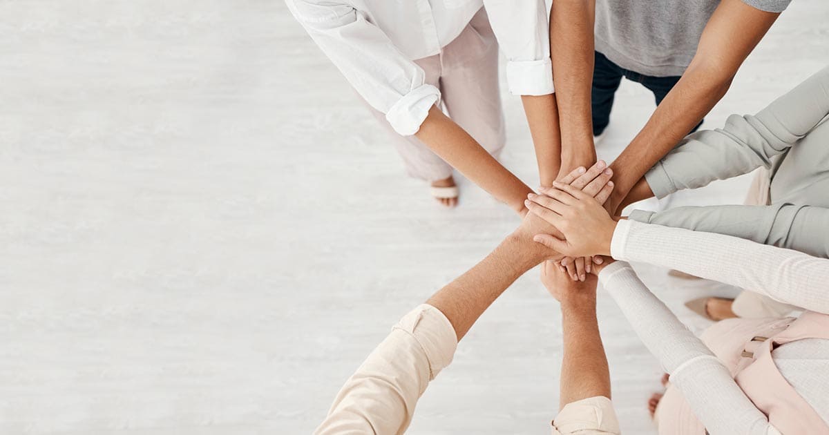 A group of people standing in a circle place their hands together in the center, symbolizing teamwork and unity—an inspiring scene often used in aspirational marketing for senior living communities.