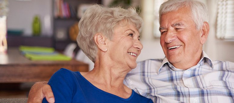 An older man and woman sit close together on a couch, smiling at each other in a warmly lit living room—an inviting scene ideal for senior living sales promotions.