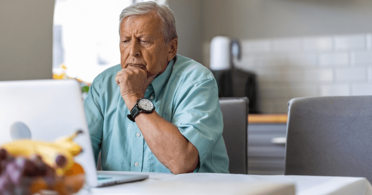An older man sits at a kitchen table, looking intently at a laptop screen with his hand on his chin, researching senior living leads.
