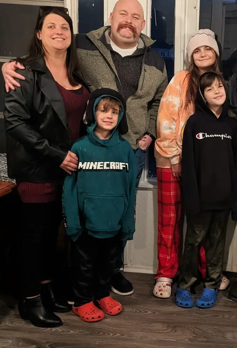 A family of five stands indoors, posing for a photo. Two adults are in the back, with three children in front, all wearing casual clothes and smiling at the camera.