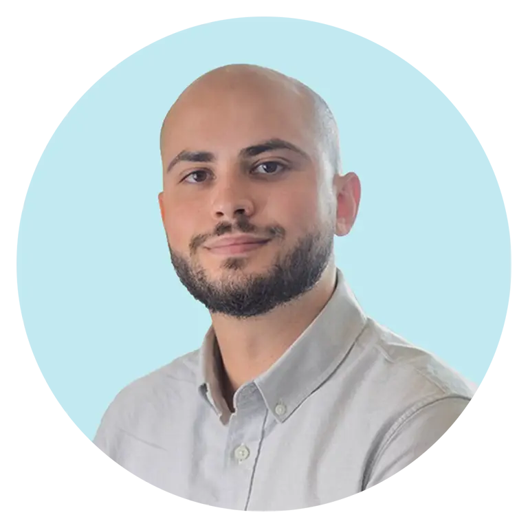 A man with a shaved head and short beard, wearing a light gray button-up shirt, poses against a light blue background.