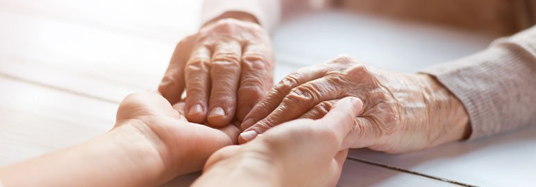 Close-up of two elderly hands resting on top of two younger hands on a white wooden surface, symbolizing comfort and support in dementia care.