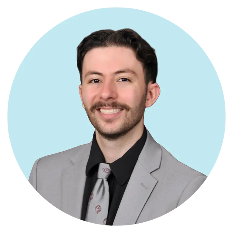 A man with short dark hair and a mustache, wearing a light gray suit, black shirt, and patterned tie, smiles at the camera against a light blue background.