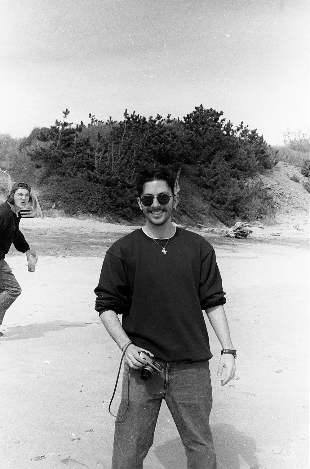 A man wearing sunglasses and holding a camera stands on a beach, smiling at the camera, while another person appears in the background making a playful expression.