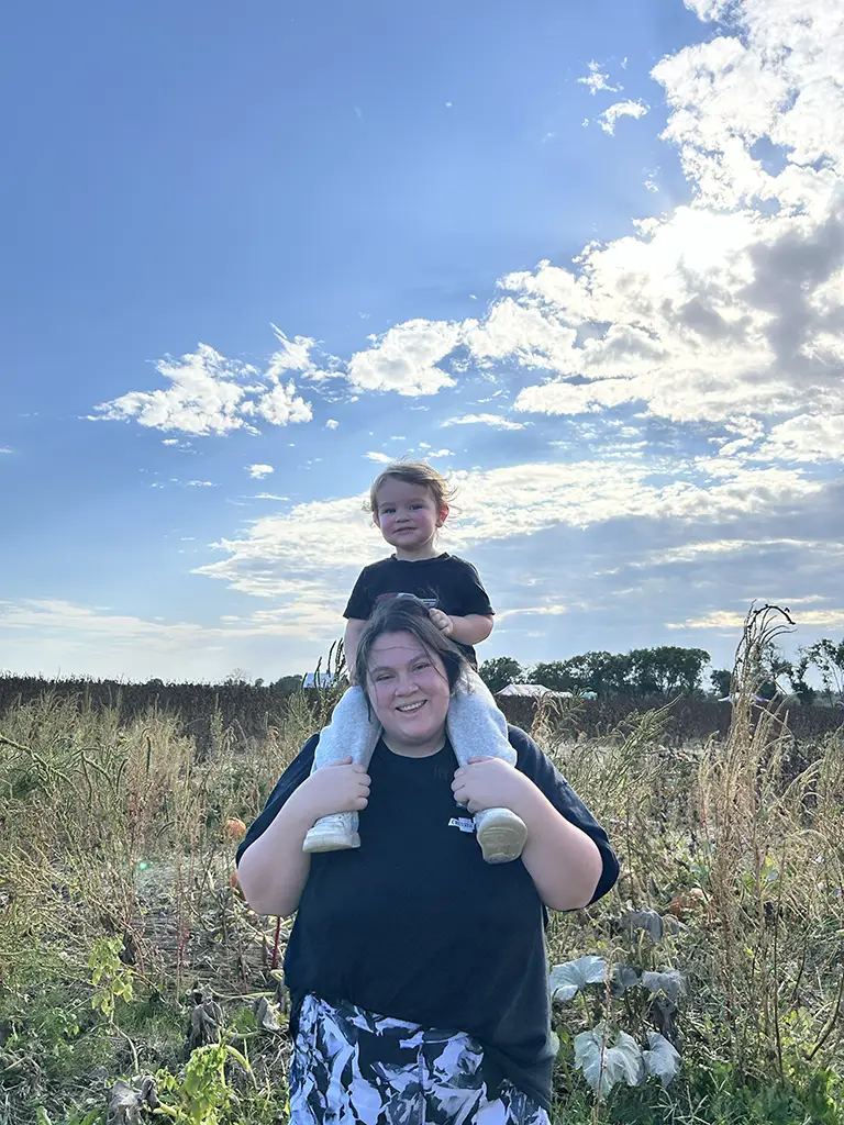An adult carries a young child on their shoulders in a sunny, grassy field with blue sky and scattered clouds in the background.