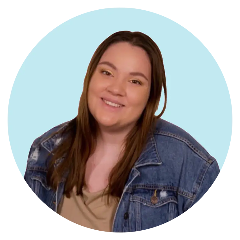 A woman with long brown hair smiles at the camera, wearing a beige top and a blue denim jacket, against a light blue background.