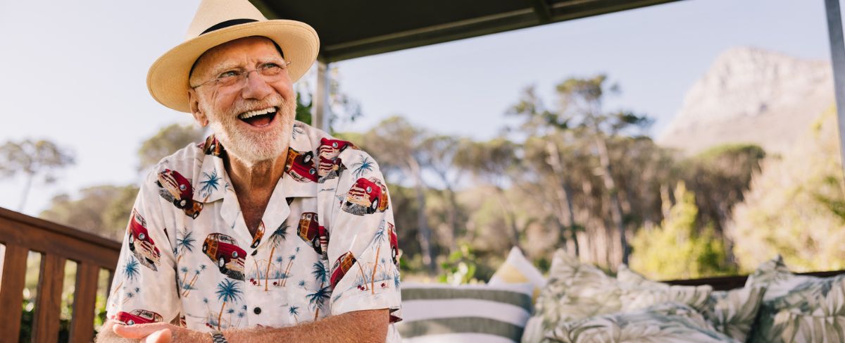 Older man wearing a straw hat and printed shirt sits outdoors, smiling and laughing, with trees, pillows, and a mountain in the background—an ideal moment for authentic baby boomer marketing.