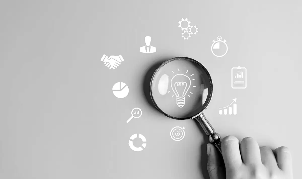 A hand holding a magnifying glass focuses on a lightbulb icon, surrounded by various business-related icons on a gray background.