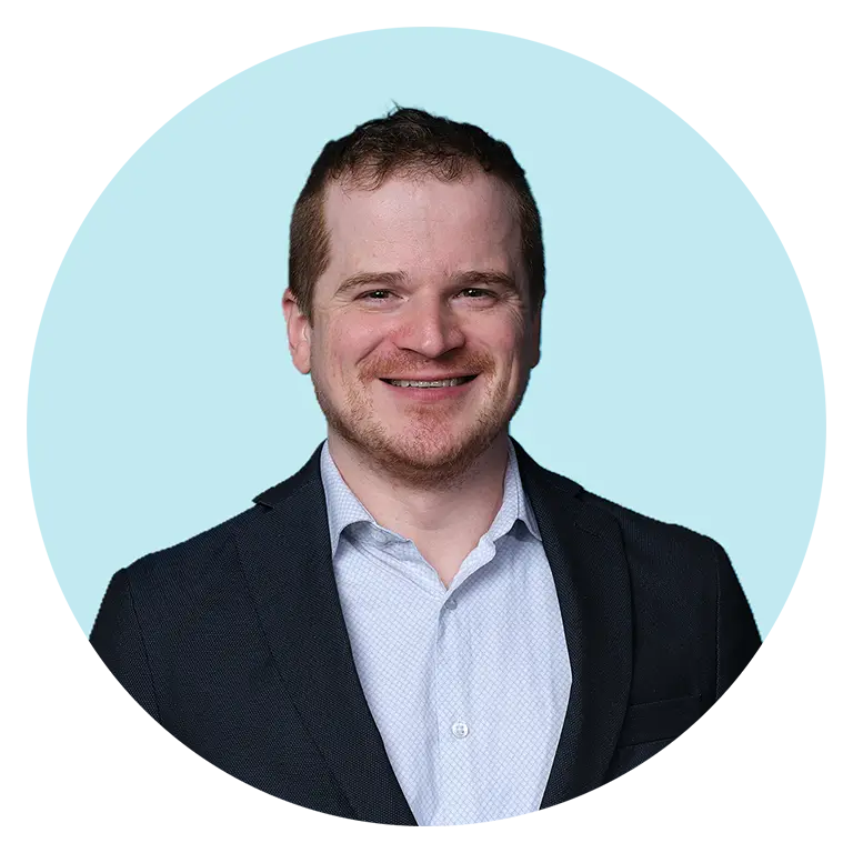 Man with short hair wearing a dark blazer and light blue dress shirt, smiling in front of a light blue background.
