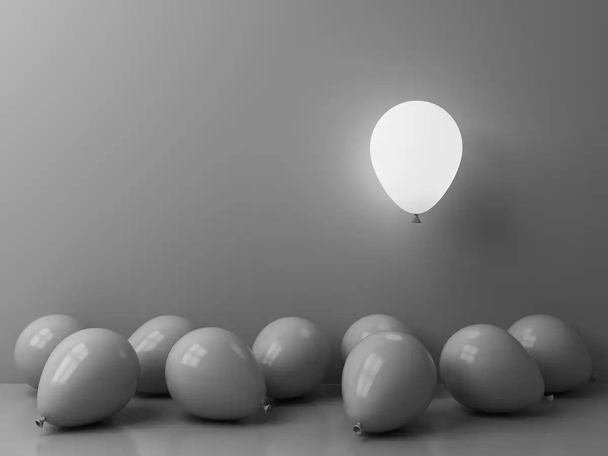 A group of balloons lies on the floor while one inflated balloon floats against a plain wall.