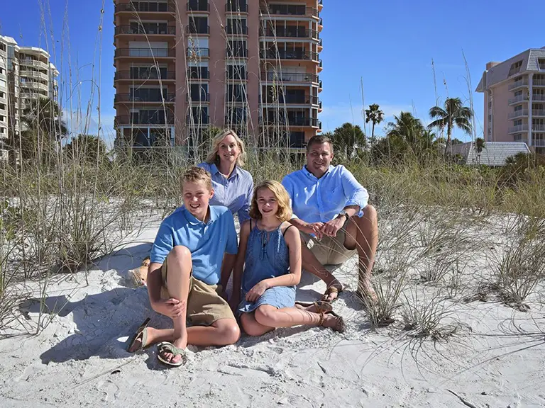 Four people, two adults and two children, sit and kneel on a sandy beach with tall grass. Apartment buildings and palm trees are visible in the background under a clear blue sky.