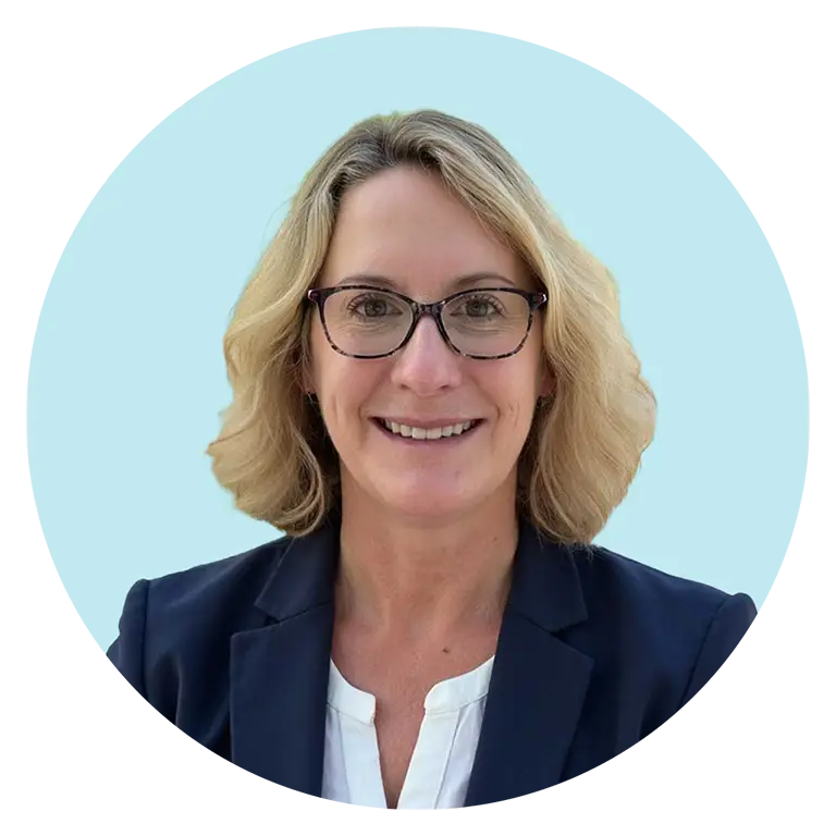 A woman with blonde hair and glasses wearing a navy blazer and white blouse, smiling at the camera against a light blue background.