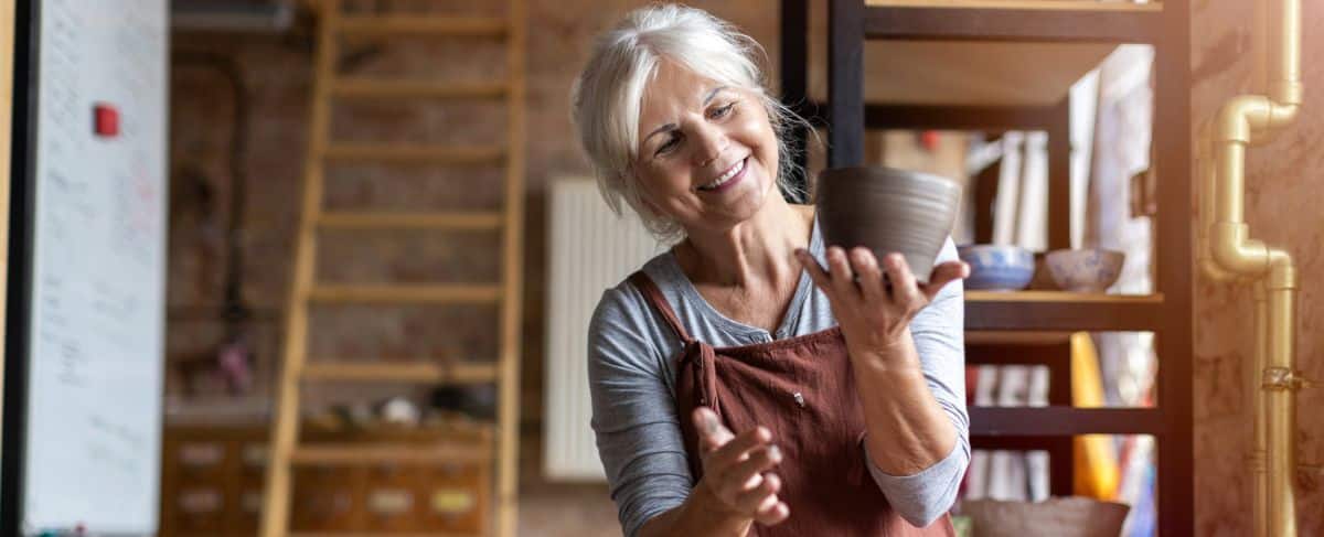 An older woman wearing an apron holds and examines a clay pot in a pottery studio, smiling as she works—capturing the creativity and engagement promoted in senior living marketing.