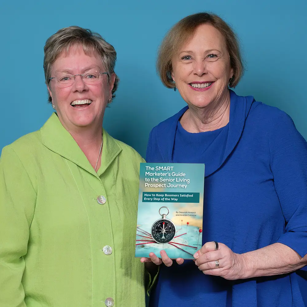 Deb-and-Andrea_Holding-Book2-1 Two smiling women stand in front of a blue background, with one holding a book titled "The SMART Marketer’s Guide to the Senior Living Prospect Journey.