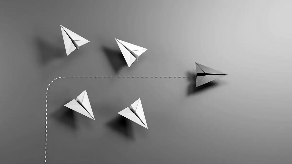 Five paper airplanes on a grey surface; four white planes fly straight, while one black plane veers off on a marked, curved path.