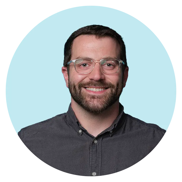 Man with short brown hair, beard, and glasses wearing a dark button-up shirt, smiling at the camera with a light blue background.