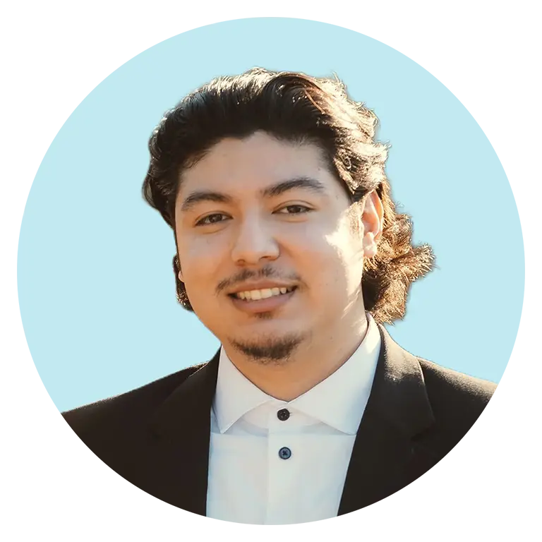 A man with medium-length dark hair and facial hair, wearing a black suit jacket and white shirt, smiles at the camera against a light blue background.