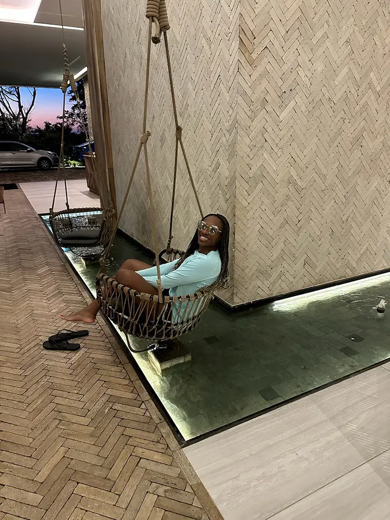 A person wearing sunglasses and a light blue outfit sits on a hanging chair over a shallow pool indoors, with sandals placed nearby on a herringbone-patterned floor.