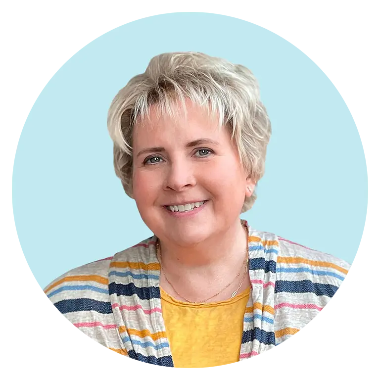 A middle-aged woman with short, light blonde hair smiles at the camera, wearing a yellow shirt and a striped cardigan, against a light blue background.