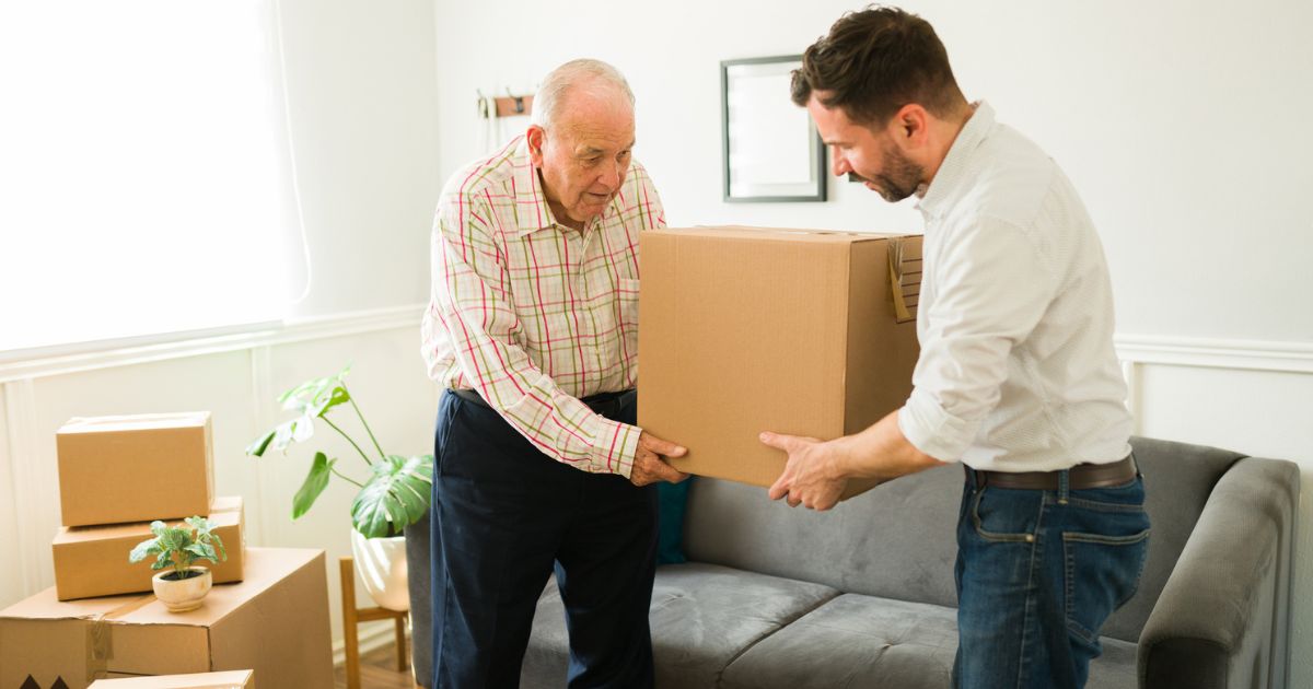 An older man and a younger man lift a cardboard box together in a living room with several packed boxes and a plant nearby, preparing for senior living tours.