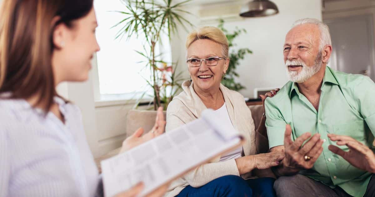 A young woman holding a clipboard speaks with an older couple sitting together on a couch in a bright, indoor setting, guiding them through the senior living sales process.