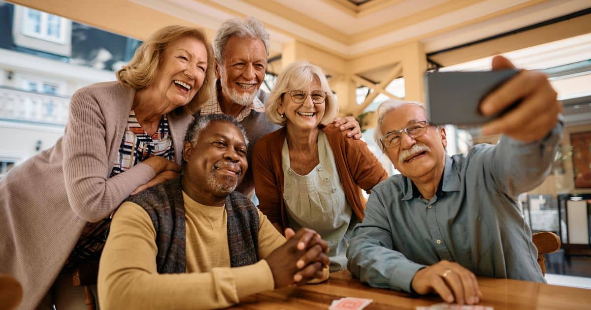 Five older adults smile while posing for a selfie together indoors, with playing cards visible on the table—a joyful moment that reflects how to create a brand experience centered around authentic connection and shared fun.