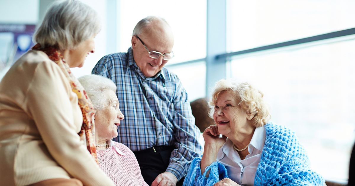 Four elderly people enjoy each other's company indoors, smiling and conversing. The spacious room's large windows in the background create a bright atmosphere, highlighting the warm sense of occupancy.
