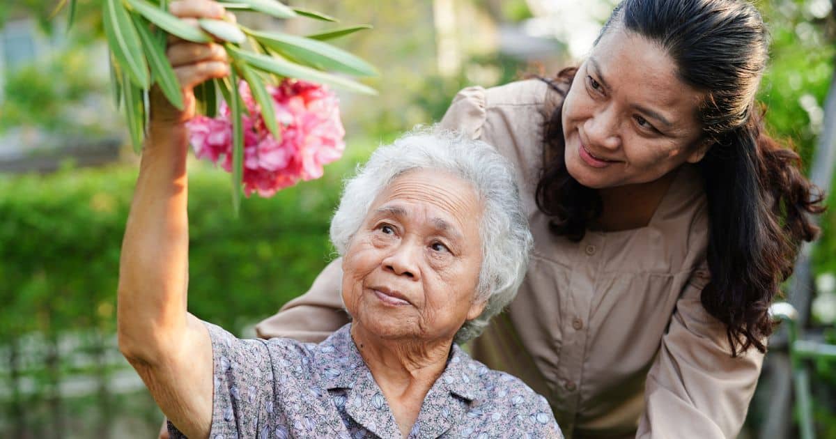 An elderly woman holds up pink flowers while a younger woman stands behind her in a lush garden—capturing a joyful moment perfect for senior living marketing.