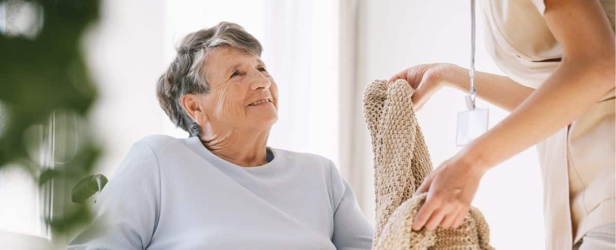 An elderly woman sitting in a wheelchair smiles up at a caregiver handing her a knitted blanket in a well-lit IL/AL community room.