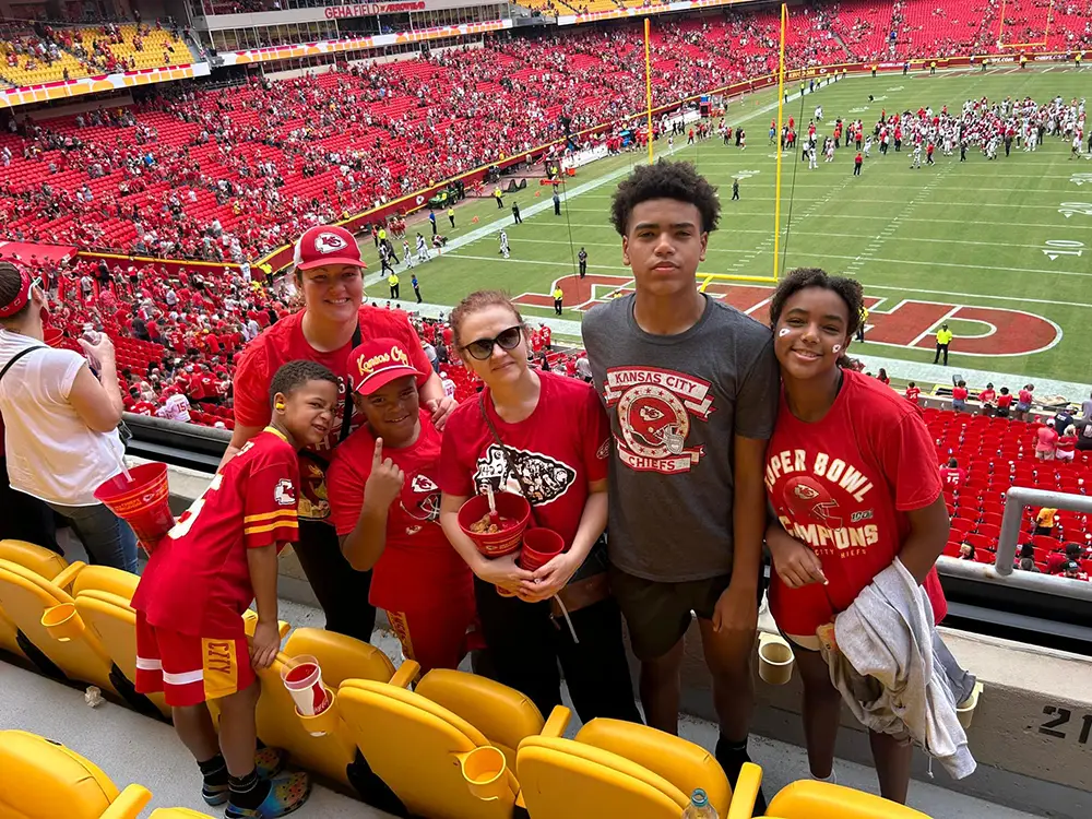 A group of six people, mostly wearing Kansas City Chiefs gear, pose for a photo in stadium seats with a football field and crowd in the background.