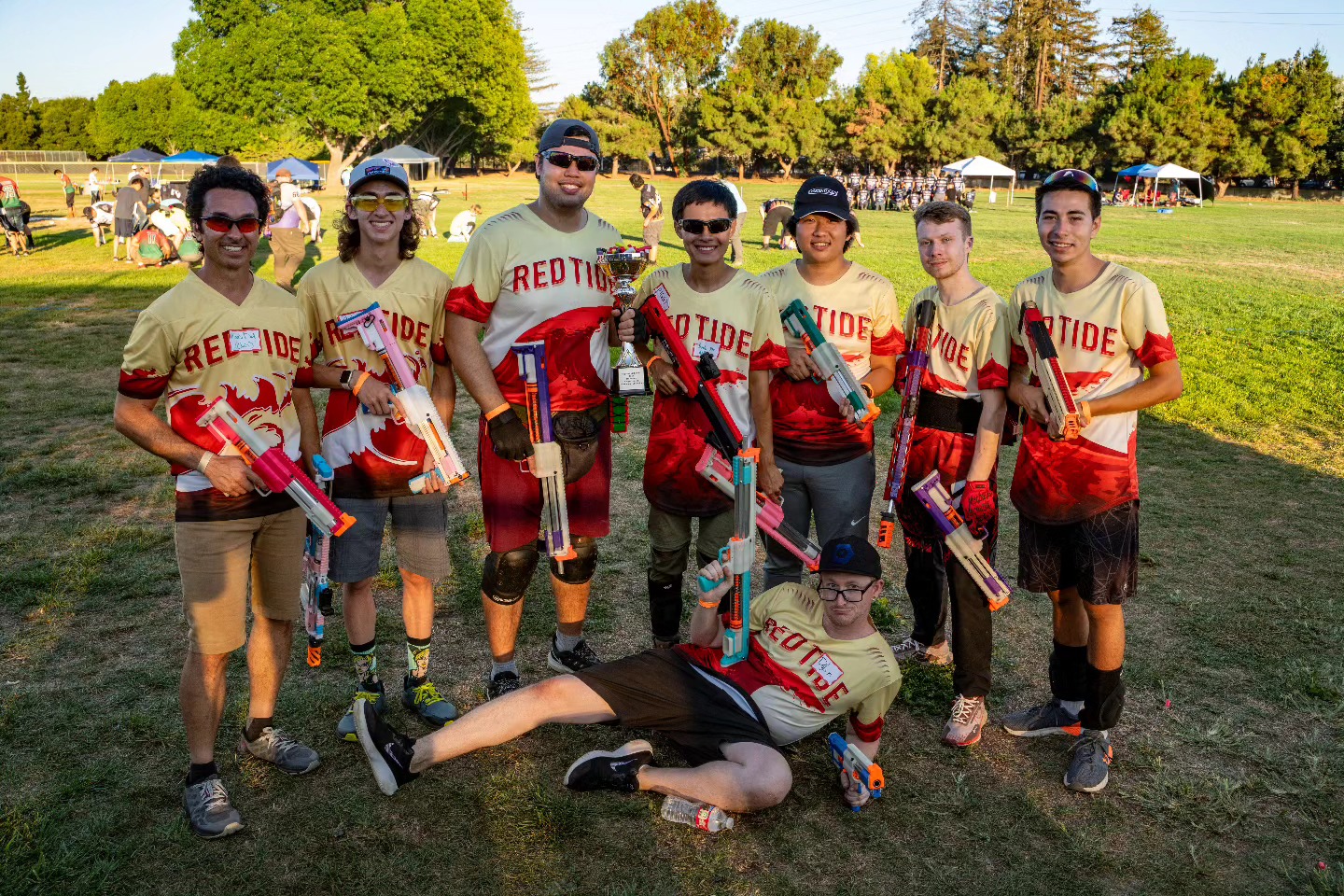 Eight people wearing matching "Red Tide" jerseys pose outdoors with toy blasters, standing on grass with trees and tents in the background. One person is lying in front of the group.
