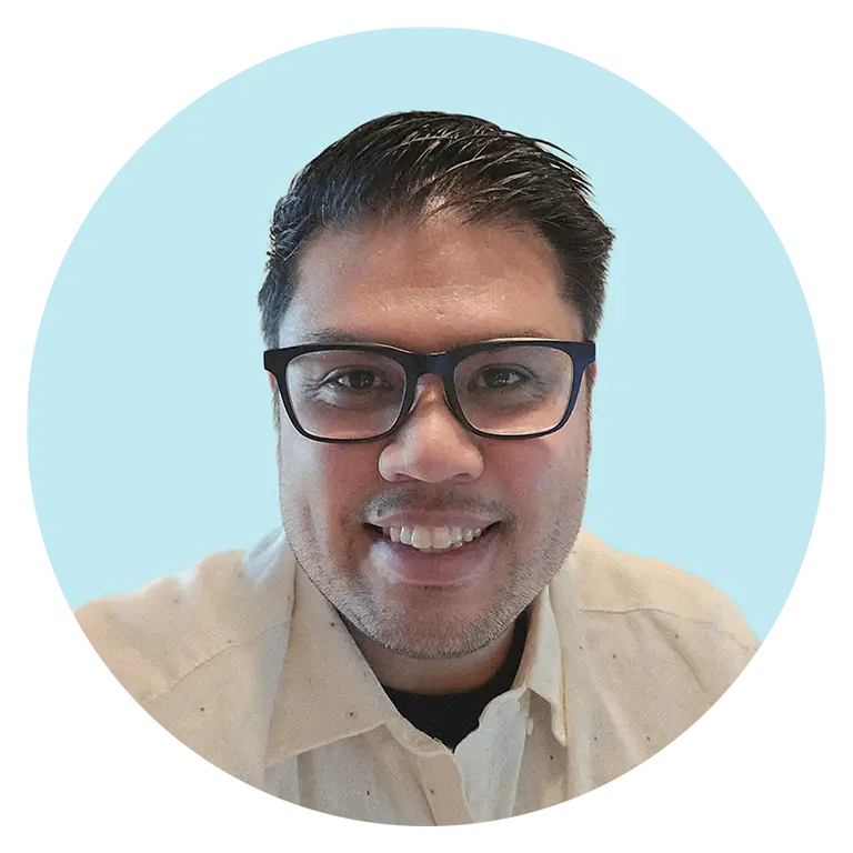 A man with dark hair and glasses smiles at the camera, wearing a light-colored shirt, against a light blue background.