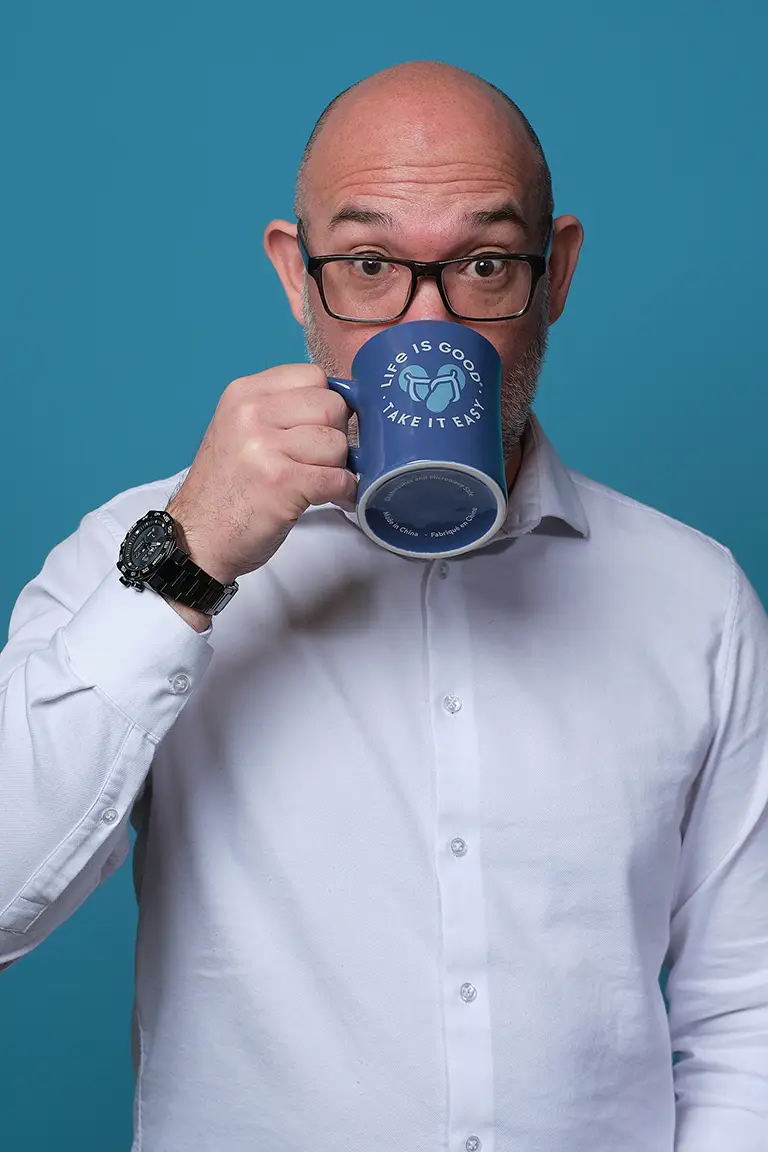 A bald man with glasses drinks from a blue mug that reads "Life is good. Take it easy." He is wearing a white shirt and a black watch, standing against a blue background.