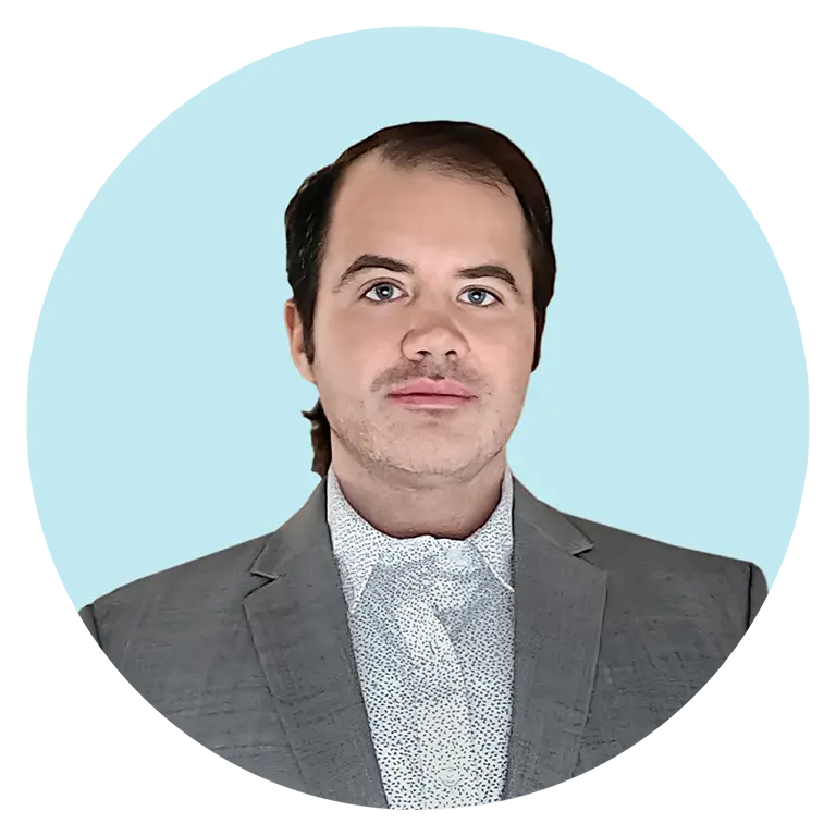 A man in a gray suit jacket and patterned white shirt stands in front of a light blue background.