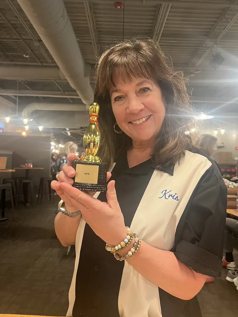 A woman wearing a black and white bowling shirt labeled "Kris" smiles while holding a small bowling trophy inside a bowling alley.