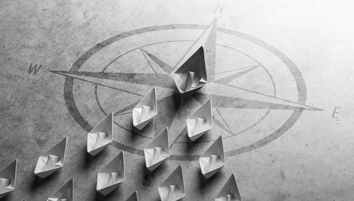 Several paper boats are arranged on a surface with a compass design, with one boat leading the others and placed at the center tip of the compass.