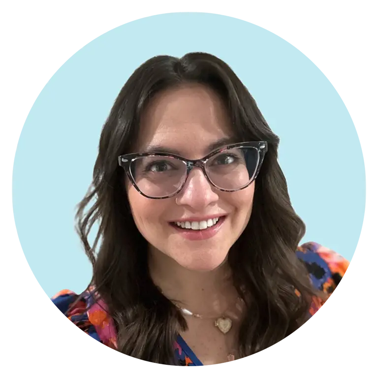 Woman with long dark hair and glasses smiling at the camera, wearing a colorful top, in front of a light blue background.