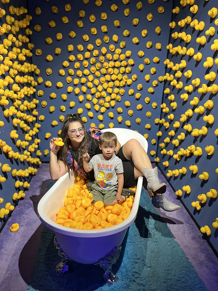 A woman and a child sit in a bathtub filled with rubber ducks, surrounded by rubber ducks attached to the blue walls of the room.