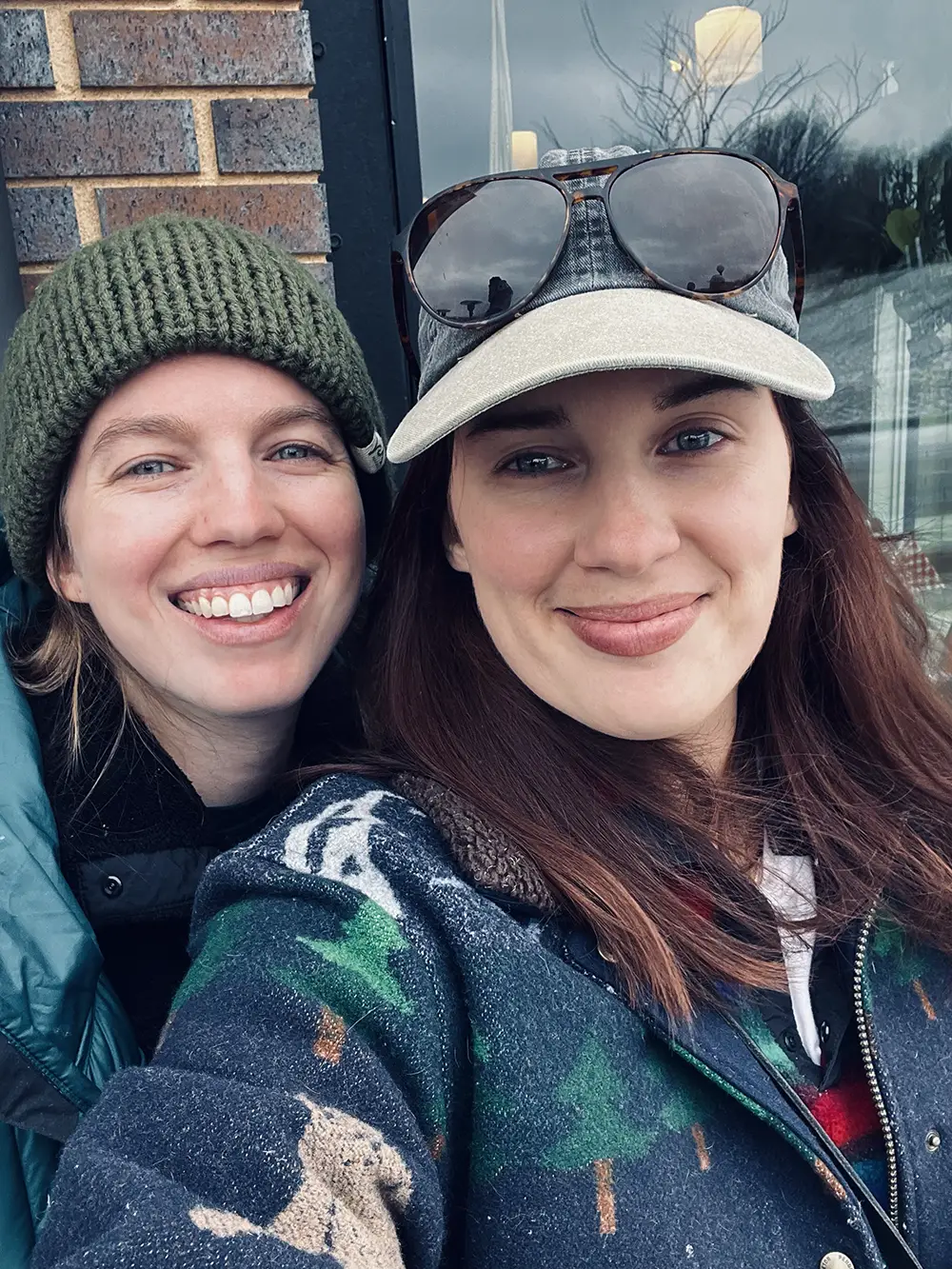 Two women smiling for a selfie outdoors; one wears a green knit hat and teal jacket, the other wears a gray baseball cap and sunglasses on her head, with a patterned jacket.