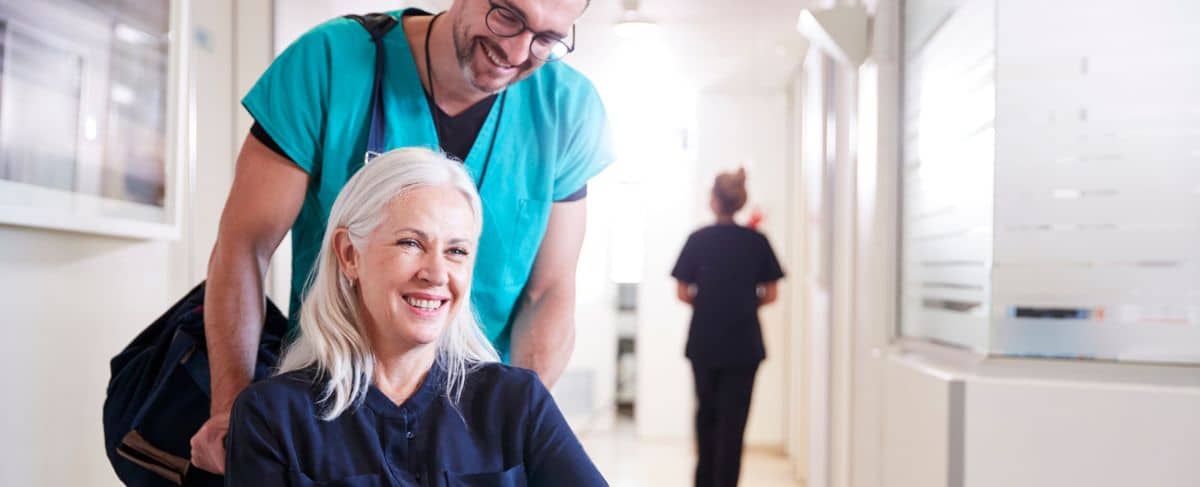 A healthcare worker in scrubs pushes a smiling older woman in a wheelchair down a hallway, demonstrating the impact of caring staff on successful retention marketing strategies; another staff member walks ahead in the background.