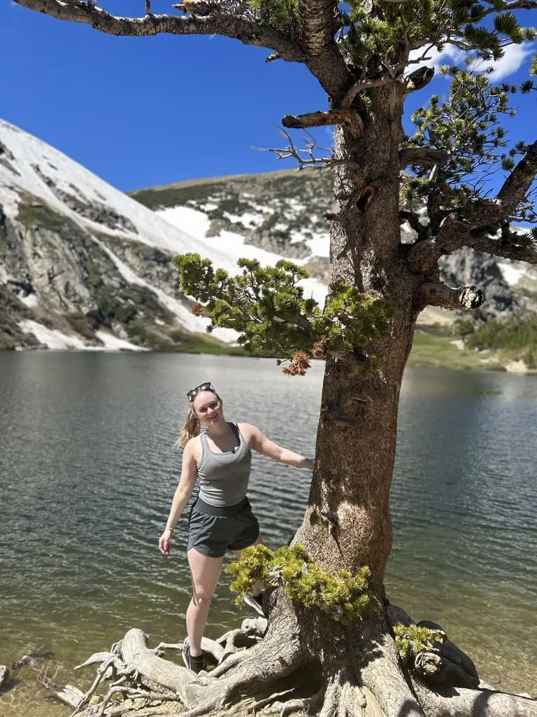 A woman stands by a large tree at the edge of a mountain lake with snow-covered slopes in the background under a clear blue sky.