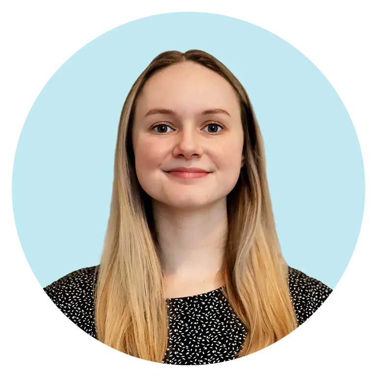 A woman with long straight blonde hair and a black patterned top smiles at the camera against a light blue background.