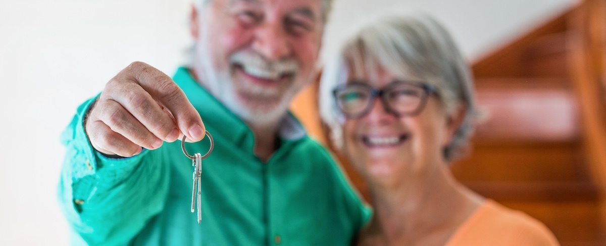 An older couple stands indoors, smiling warmly, as one person holds a set of keys toward the camera—capturing the excitement and promise of senior living sales marketing.