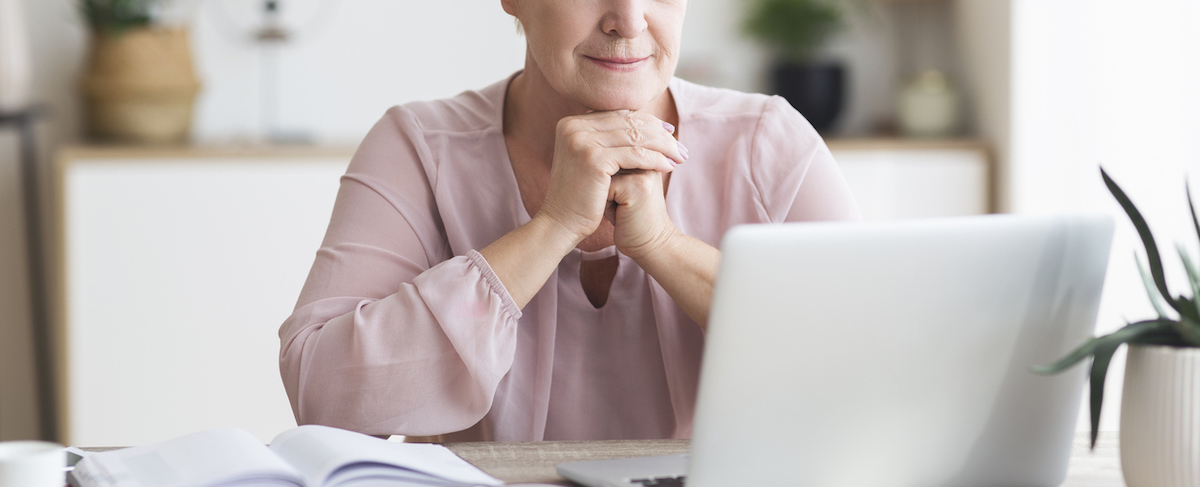 A person sits at a desk with hands clasped, looking at a laptop screen, with an open notebook and pen nearby, thoughtfully reviewing a senior living marketing strategy.