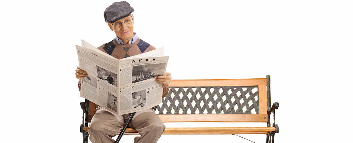 An older man wearing a cap and glasses sits on a wooden bench, reading a newspaper against a plain white background—an ideal image for senior living advertising.